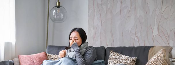Женщина с температурой, кашлем, симптомы гриппа, гриппа или холода, сидит в теплой одежде на диване, покрытый одеялом, чихает, остается дома в гостиной. - Фото, изображение