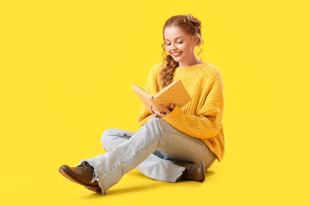Gyönyörű boldog fiatal nő meleg pulóverben ül a sárga háttér és olvasás könyv - Fotó, kép