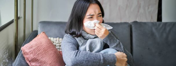 Primer plano de chica coreana estornudando en siesta, sentado en casa en bufanda, sintiéndose mal, concepto de invierno, costo de calefacción y personas. - Foto, imagen