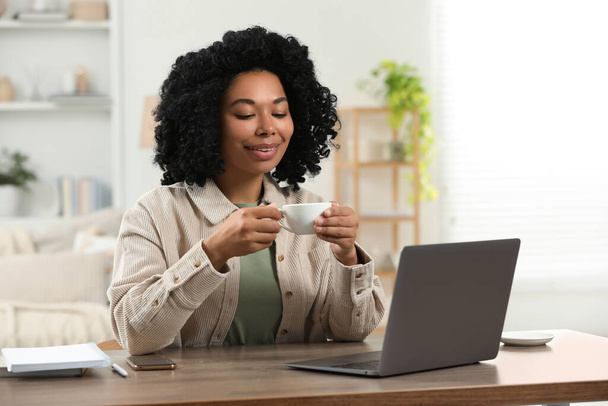 Happy young woman with cup of drink using laptop at wooden desk indoors - Фото, изображение
