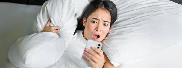 Közelkép portré ázsiai lány feküdt az ágyban, nézi okostelefon érintett, felébredt későn, és bámulta az ébresztőórát a mobiltelefon. - Fotó, kép