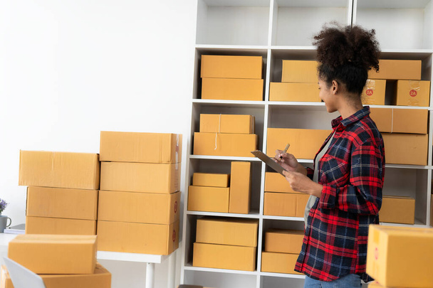 Afrykanka sprzedaje produkty online przez internet siedząc na kanapie w domu, ciesząc się chwilą z kartonowym pudełkiem z paczki, ciesząc się z dostawy. - Zdjęcie, obraz