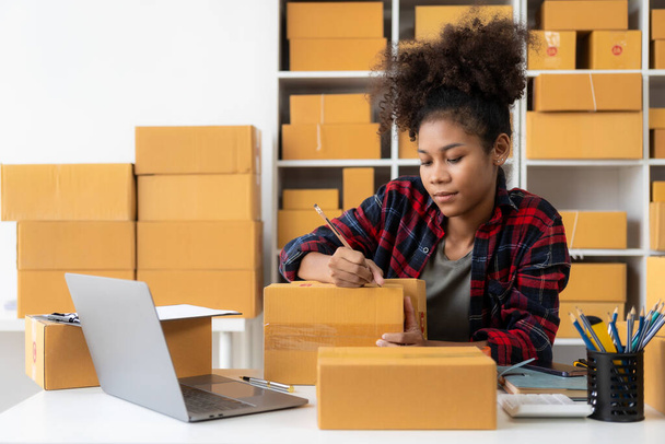 Африканская женщина, продающая онлайн продукты через интернет сидя на диване дома, наслаждаясь моментом с картонной коробкой из посылки, чувствуя себя счастливым от доставки. - Фото, изображение