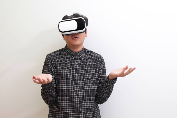 Retrato de un hombre asiático con camisa a cuadros negra usando gafas de Realidad Virtual (VR) y tratando de sostener algo frente a él. Imagen aislada sobre fondo blanco - Foto, imagen