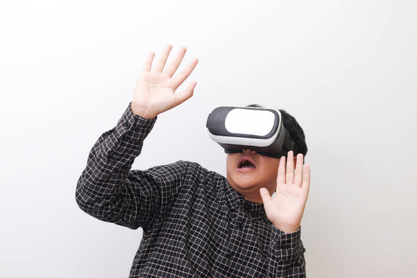 Retrato del hombre asiático con camisa a cuadros negra usando gafas de Realidad Virtual (VR) y protegiéndose extendiendo sus brazos desde algo grande cayendo desde arriba. Imagen aislada sobre fondo blanco - Foto, imagen