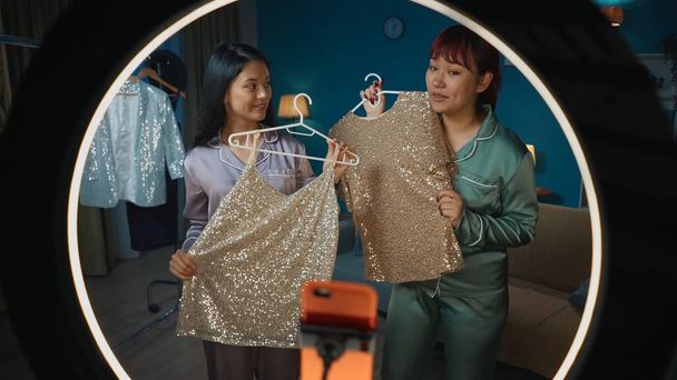 Full-sized photo shot from inside the ring light capturing two young women filming themselves on a smartphone, sharing live as they show different clothes they have on a hanger. Fashion blogger. - Zdjęcie, obraz