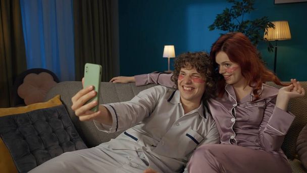 Beauty and healthy relationships advertisement concept. Portrait of young couple spending time together. Man and woman sitting on the sofa, wearing pajamas and eyepatches, recording selfie video. - 写真・画像