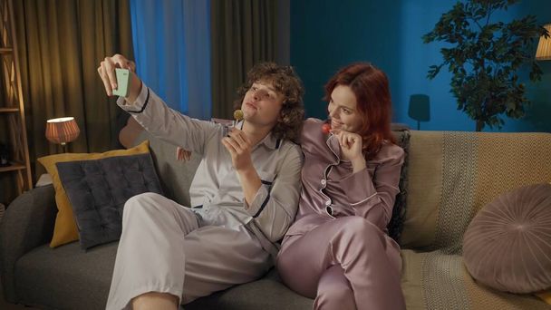 Beauty and healthy relationships advertisement concept. Portrait of young couple. Man and woman in pajamas sitting on the sofa eating lollipops, taking selfie on smartphone. - Photo, Image