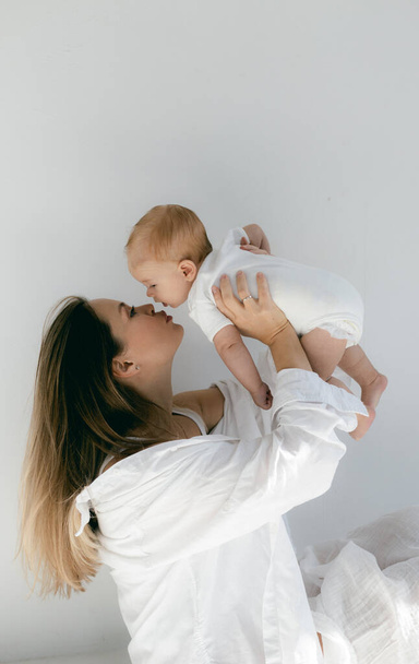 Jonge vrouw met baby in haar armen. Blond meisje in witte kleren glimlachend naar uw kind op witte achtergrond. Gelukkig moederschap en borstvoeding concept. Foto 's met zonneschijn, zachte focus, overbelichting. - Foto, afbeelding