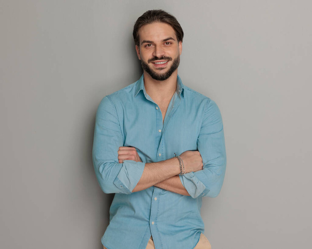 attraktiver junger Mann mit verschränkten Armen, lächelnd und posierend vor grauem Hintergrund  - Foto, Bild