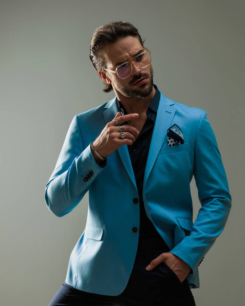 Mode eleganter Mann mit Brille, der zur Seite schaut, während er mit der Hand in der Tasche vor grauem Hintergrund posiert - Foto, Bild