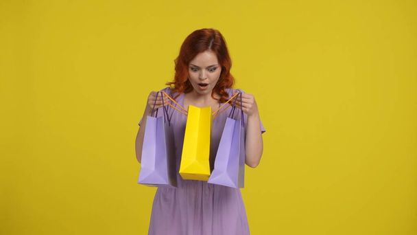 A young redhaired woman looks into her shopping bag with surprise. Woman in the studio on a yellow background. Sale, Black Friday, advertising, discounts - Фото, зображення