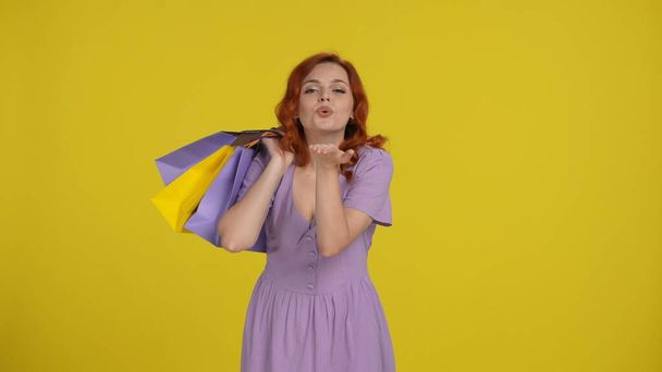 Satisfied redhaired woman with shopping bags blows a kiss. Woman in the studio on a yellow background. Sale, Black Friday, advertising, discounts - Фото, зображення