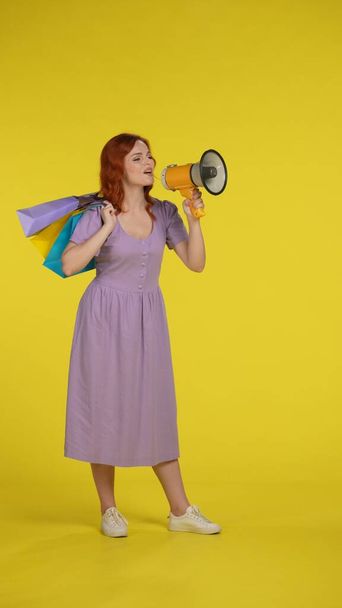 Happy redhaired woman with shopping bags speaking into a megaphone. Full length woman in the studio on a yellow background. Vertical shot. Sale and discount concept - 写真・画像