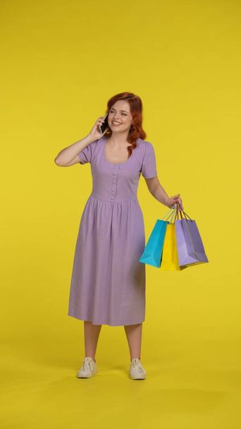 A woman with shopping bags speaks on a smartphone, shares her emotions from shopping. Full length woman in the studio on a yellow background. Vertical shot. Sale, Black Friday, advertising, discounts - Foto, immagini