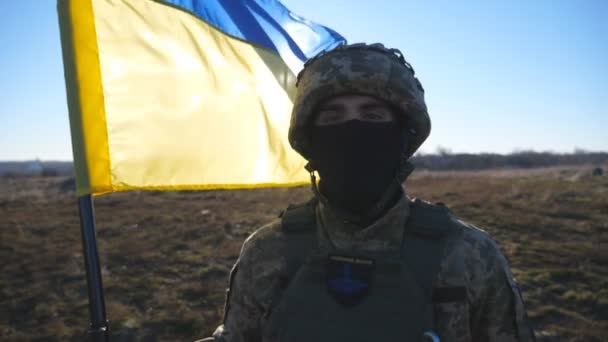 Portrait de soldat masculin en uniforme de camouflage tenant un drapeau ondulé de l'Ukraine. Homme de l'armée ukrainienne dans le casque et la cagoule avec lever bannière bleu-jaune regarde dans la caméra. invasion russe de l'Ukraine - Séquence, vidéo