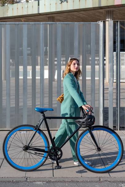 Stylová rusovlasá žena v zeleném obleku, procházka na kole ve finanční čtvrti s moderní budovou na pozadí - Fotografie, Obrázek