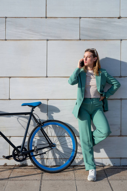 Joven mujer pelirroja con poderes en traje verde hablando en el teléfono inteligente de pie con la bicicleta en el fondo de mármol - Foto, imagen