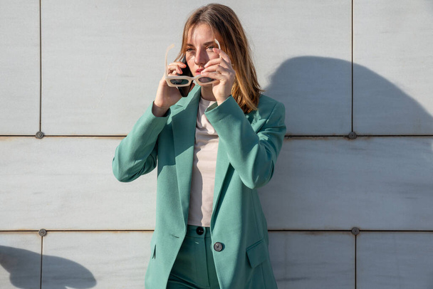 Молода жінка з імбирним волоссям у зеленому костюмі говорить на смартфоні, стоячи на мармуровому фоні - Фото, зображення