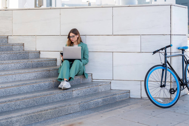 Mulher de negócios sentada em escadas fora do prédio de escritórios enquanto trabalhava com laptop ao lado de uma bicicleta - Foto, Imagem