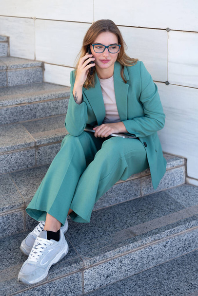 Business woman sitting on stairs outside office building while working with laptop and having a phone call - Photo, Image