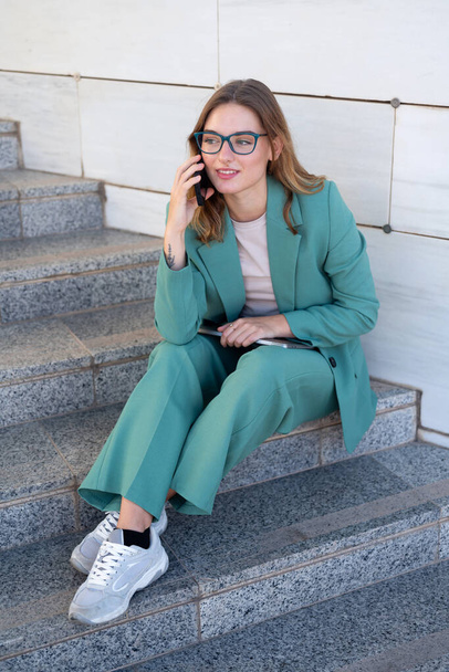 Femme d'affaires assise dans les escaliers à l'extérieur de l'immeuble de bureaux tout en travaillant avec un ordinateur portable et ayant un appel téléphonique - Photo, image