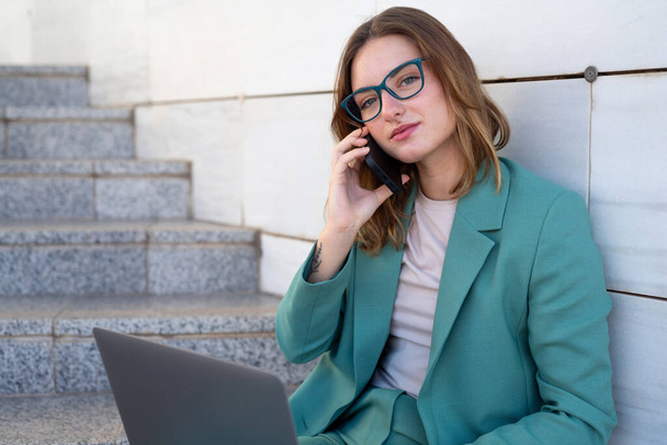 Zakelijke vrouw zit op de trap buiten kantoorgebouw tijdens het werken met laptop en het hebben van een telefoontje - Foto, afbeelding
