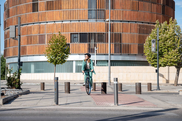 Tyylikäs liiketoiminnan nainen vihreässä puvussa ja kypärä ratsastus hänen polkupyörän rahoitusalueella moderni rakennus taustalla - Valokuva, kuva
