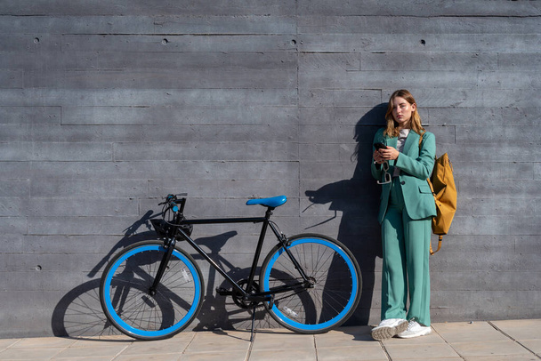 Молода сильна жінка з імбирним волоссям у зеленому костюмі, використовуючи смартфон, що стоїть зі своїм велосипедом на чорному тлі - Фото, зображення