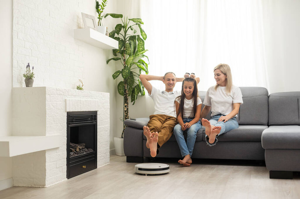 Молодая семья отдыхает на диване, в то время как роботизированный пылесос делает свою работу дома. Концепция бытовых роботов - Фото, изображение