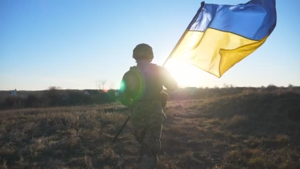 Soldat masculin de l'armée ukrainienne fonctionne avec levé bannière nationale sur le terrain. Jeune homme en uniforme de camouflage jogging avec le drapeau de l'Ukraine agitant sur prairie au coucher du soleil. Concept de résistance à l'invasion. Vue arrière. - Séquence, vidéo