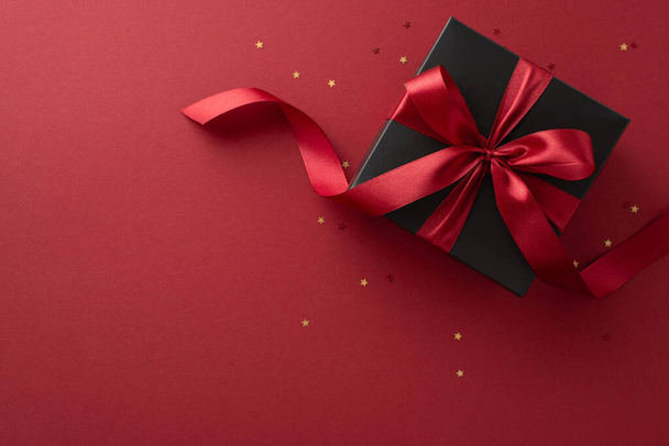 Black Friday surprise: Top view of a deluxe black gift box featuring a stylish red ribbon and gold confetti on a marsala background, offering an ideal spot for your text or ad - Photo, Image