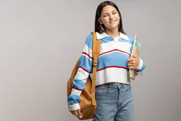 Счастливая улыбающаяся студентка-подросток с рюкзаком, копирует книги на заднем плане. Портрет брюнетка симпатичный дружелюбный подросток в толстовке, джинсах. Рекламный баннер с копировальным пространством, концепция образования. - Фото, изображение
