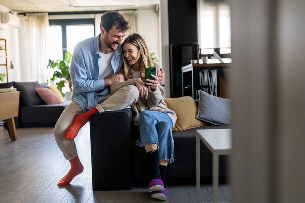 Young happy couple using phone to share social media news at home, smiling talking doing shopping online. Mobile tech lifestyle concept - Valokuva, kuva