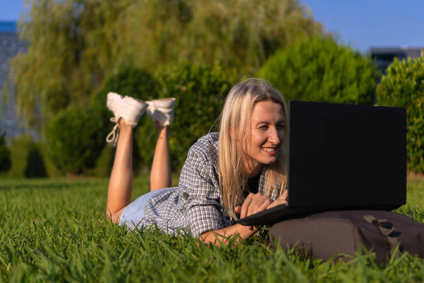Portrait of a young smiling blonde woman lying on her stomach on the grass in the park with a laptop on a sunny day. Student communicates, studies with a laptop in a city park - Photo, Image
