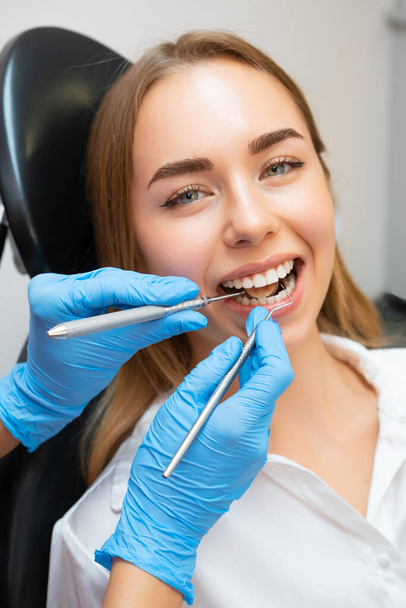歯科クリニックで歯科治療中にアームチェアに座っている閉じた女性患者. 歯の健全性をチェックする器具で前歯をチェックする医師 - 写真・画像
