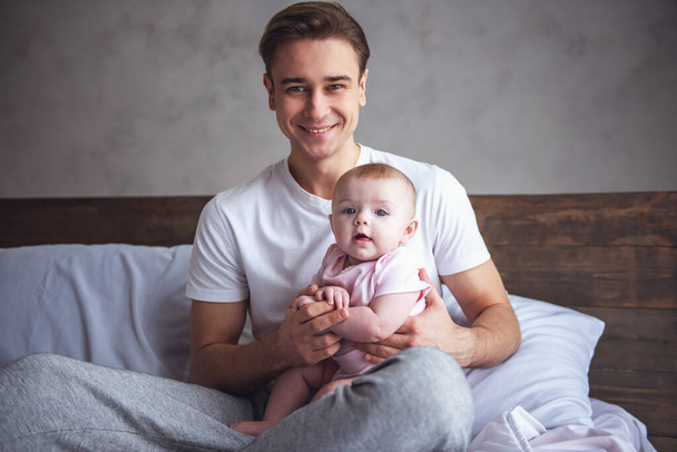 Beau jeune papa tient son bébé mignon dans les bras, regardant la caméra et souriant tout en étant assis sur le lit
 - Photo, image