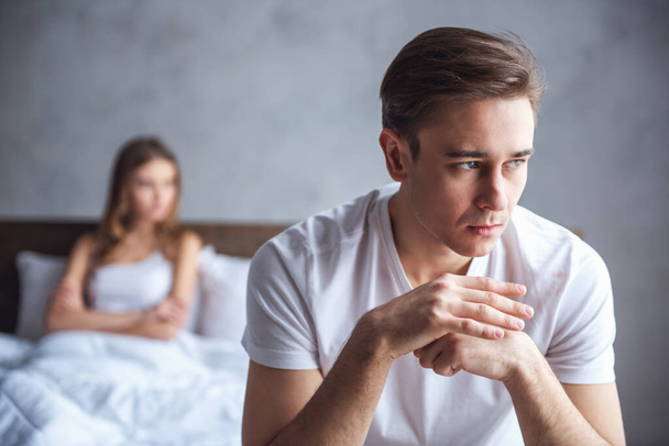 O jovem casal tem um problema. Guy está sentado na cama e olhando tristemente para longe, sua namorada no fundo
 - Foto, Imagem