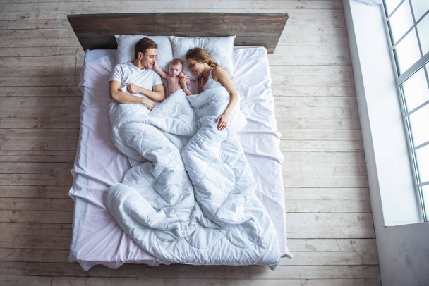 Вид сверху на красивых молодых родителей, которые смотрят на своего симпатичного ребенка и улыбаются, лежа вместе в постели
 - Фото, изображение