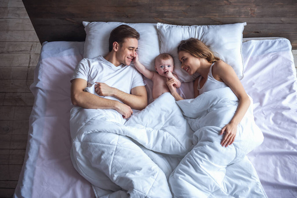 彼らはかわいい赤ちゃんを見ていると、ベッドで一緒に横になって微笑む美しいの若い親の平面図 - 写真・画像