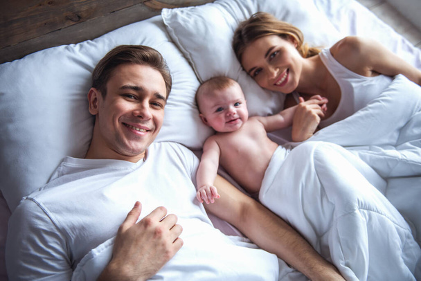 Ritratto di bei giovani genitori e bambino carino guardando la macchina fotografica e sorridendo mentre sdraiati insieme a letto
 - Foto, immagini