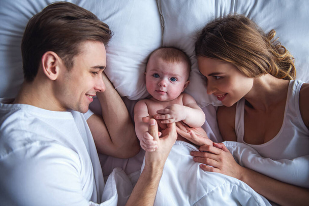 Kauniit nuoret vanhemmat katsovat söpöä vauvaansa ja hymyilevät sängyssä.
 - Valokuva, kuva