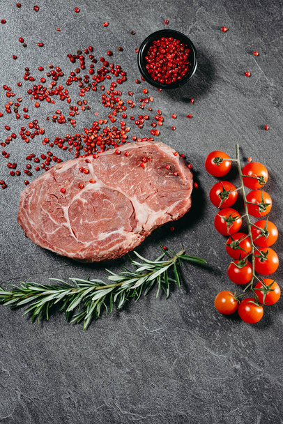 Top view flat lay on gray stone cutting table on which is a fresh steak with a sprinkle of red pepper, which is in a black saucer, sprigs of rosemary and cherry tomatoes next to a kitchen knife - Photo, Image