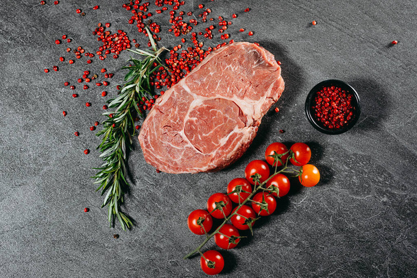 Fresh ribeye steak with red pepper sprinkles in a black saucer, rosemary sprigs and cocktail tomatoes next to a kitchen knife on a top view flat lay on gray stone cutting table - Photo, Image