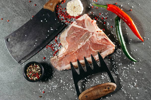 A piece of fresh bone-in ribeye steak, sprinkled with salt and red peppercorns, lying on a stone table, next to which is a wide hatchet knife, a sharp meat holder, sliced garlic, a sprig of rosemary, a black saucer with red and black peppercorns it a - Photo, Image