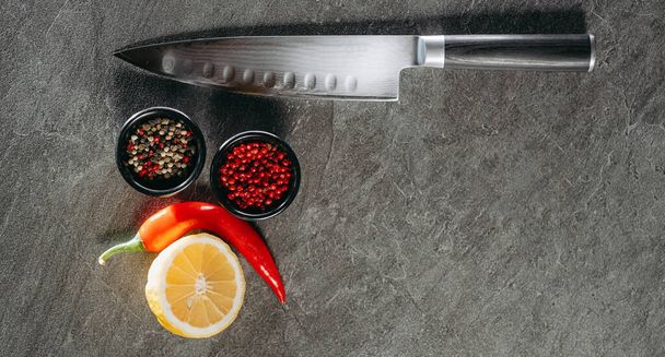 Великий гострий кухонний ніж з чорною ручкою, поруч дві чорні чашки з червоним перцем і міксом, гарячий червоний перець і нарізаний шматочок лимона, які знаходяться на кухонному кам'яному столі - Фото, зображення