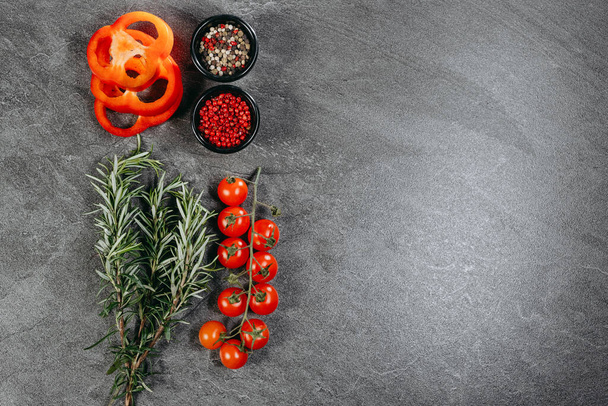 Estetyczny układ gałązek pachnącego rozmarynu, trzy pierścienie czerwonej papryki, gałąź pomidorów wiśniowych i dwa czarne kubki z mieszanym groszkiem pieprzowym i czerwonym na kamiennym stole kuchennym - Zdjęcie, obraz