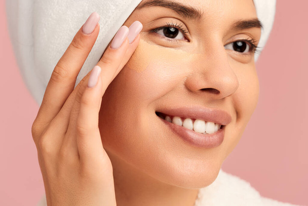 Young happy female model in bathrobe with towel on head applying concealer under eyes with fingers while enjoying beauty procedure against pink background - Foto, imagen