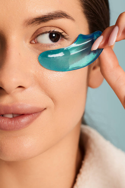Crop macro shot of a face of young female applying blue eye patches on blue background during skin care routine - Fotografie, Obrázek