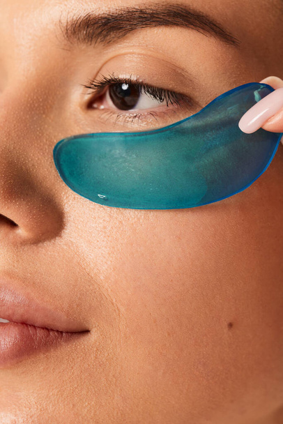 Closeup of crop anonymous female looking away while applying under eye silicone gel moisturizing patch with manicured fingers during skincare procedure in light - 写真・画像